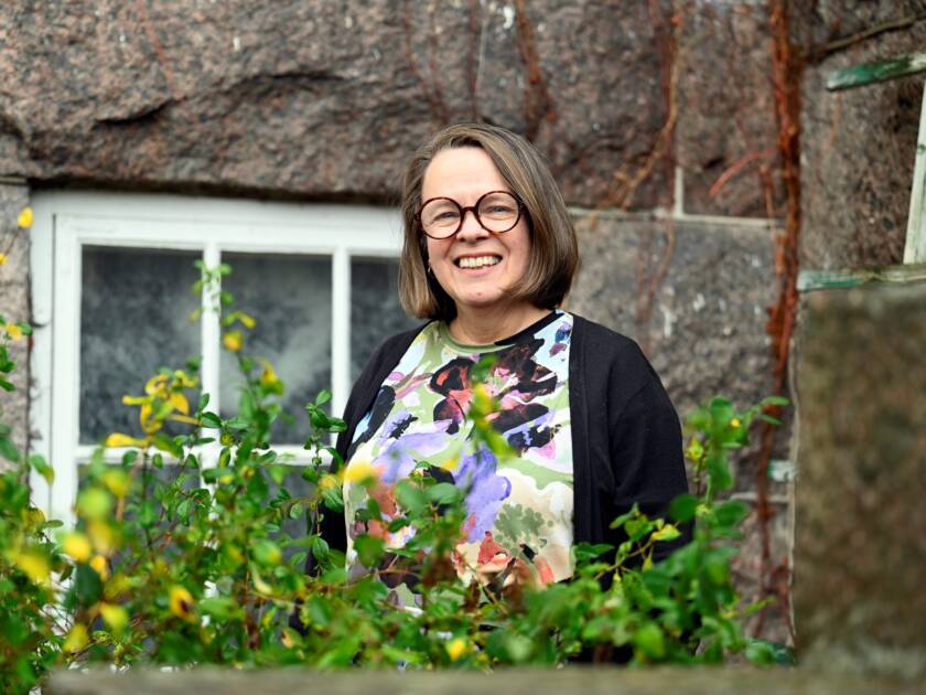 hymyilevä nainen taustalla kiviseinä, etualalla kukkia
