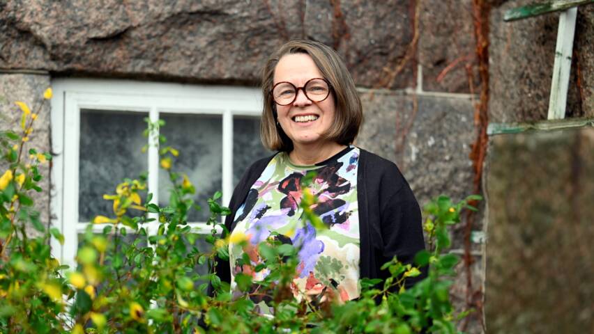 hymyilevä nainen taustalla kiviseinä, etualalla kukkia