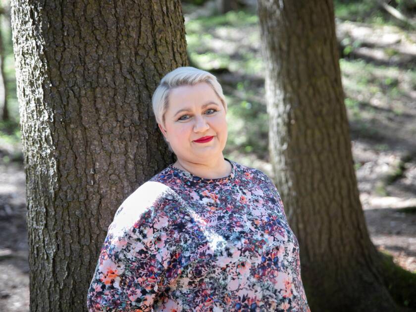 Kuvassa vaalea lyhythiukisnen nainen nojaa puunrunkoon