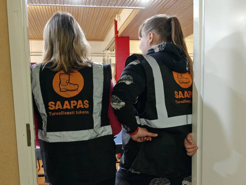 Kuvassa kaksi ihmistä selin kameraan päin. Selässä teksti Saapas - turvallisesti tukena.